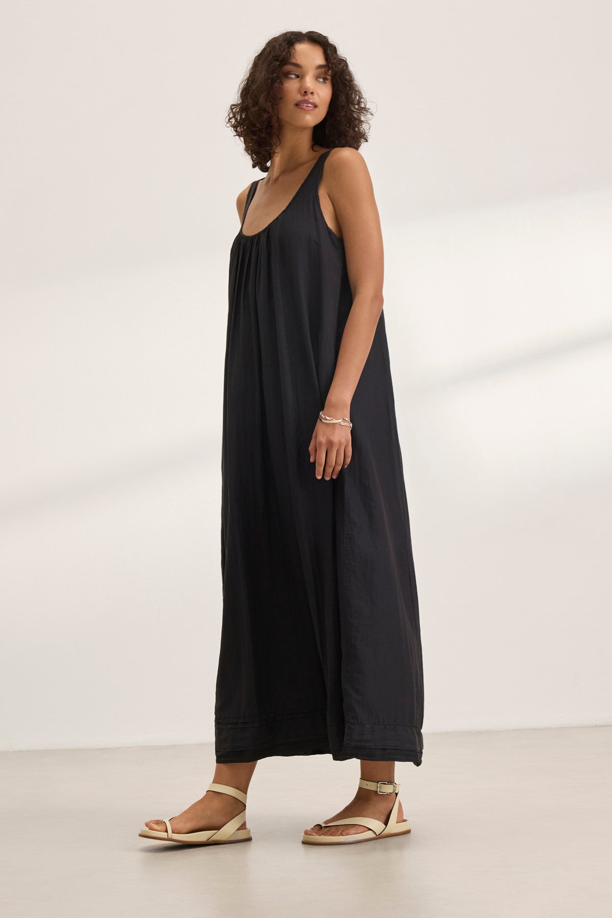 In a neutral backdrop, a person wearing the BAYOU SILK COTTON VOILE DRESS by Velvet by Graham & Spencer gazes slightly left. The sleeveless, long black maxi dress elegantly drapes, paired with sandals and a bracelet for versatile styling.-38801130979521