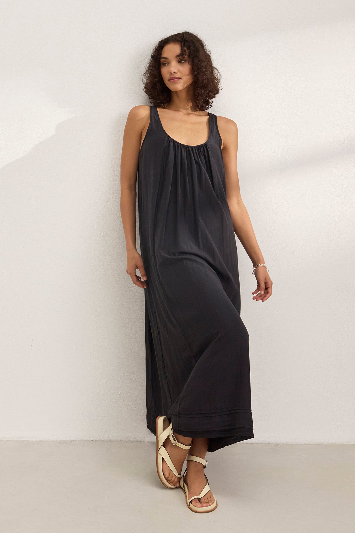 A woman wears the BAYOU SILK COTTON VOILE DRESS by Velvet by Graham & Spencer—a sleeveless black maxi in silk cotton voile—with white sandals, standing against a plain white wall.-39273978298561