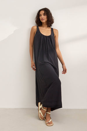A woman wears the BAYOU SILK COTTON VOILE DRESS by Velvet by Graham & Spencer—a sleeveless black maxi in silk cotton voile—with white sandals, standing against a plain white wall.