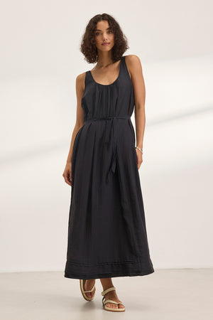 A woman stands indoors wearing the BAYOU Silk Cotton Voile Dress by Velvet by Graham & Spencer—an ankle-length, sleeveless black dress with a scoop neck, tie belt, and effortless drape—paired with flat sandals.