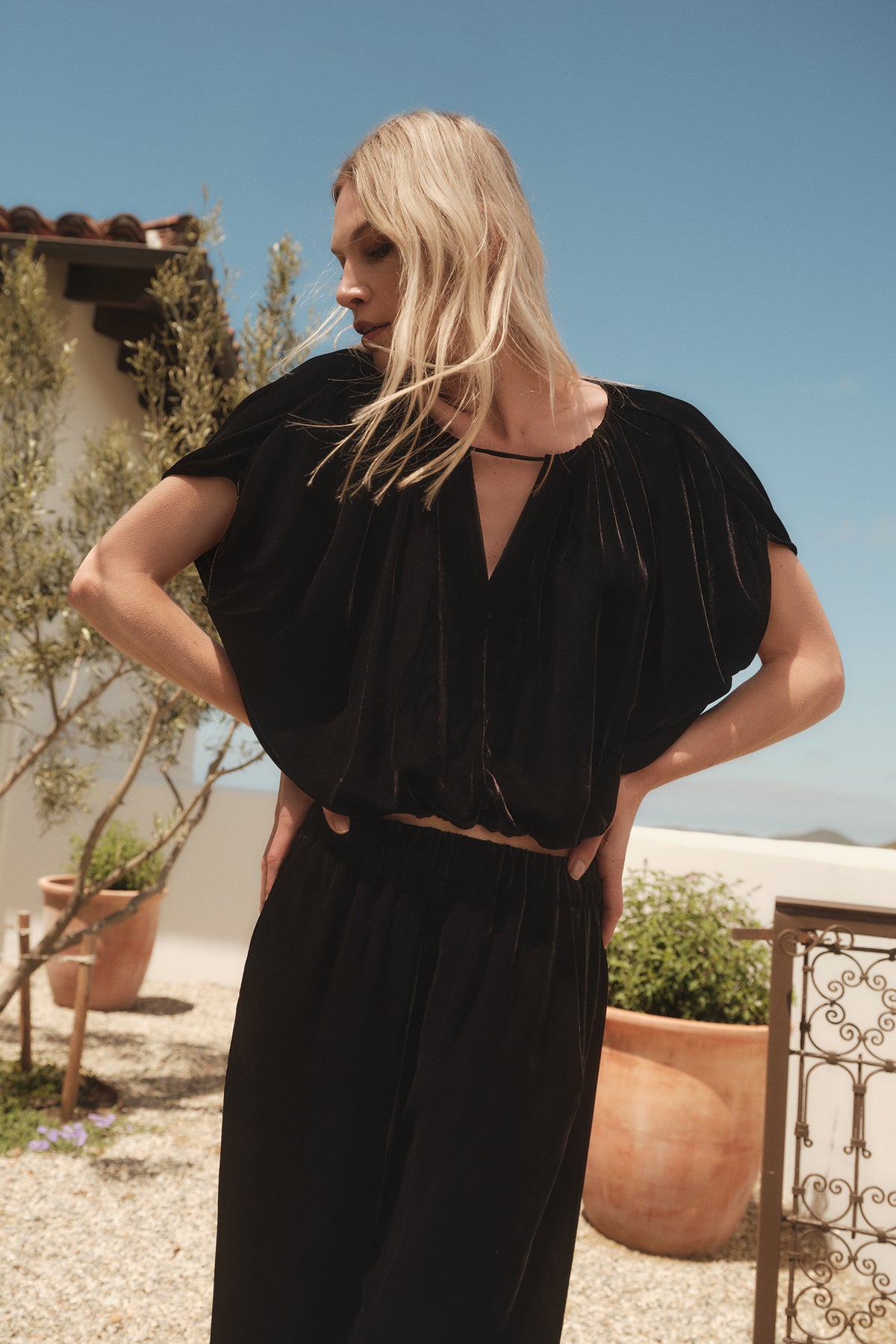 A blonde woman wearing a black NOA SILK VELVET CROPPED TOP jumpsuit by Velvet by Graham & Spencer with an adjustable tie and V-neck top.-35654468731073