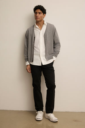 A man stands against a plain wall, wearing a white shirt, black pants, white sneakers, and the SILVIO CARDIGAN in gray by Velvet by Graham & Spencer.