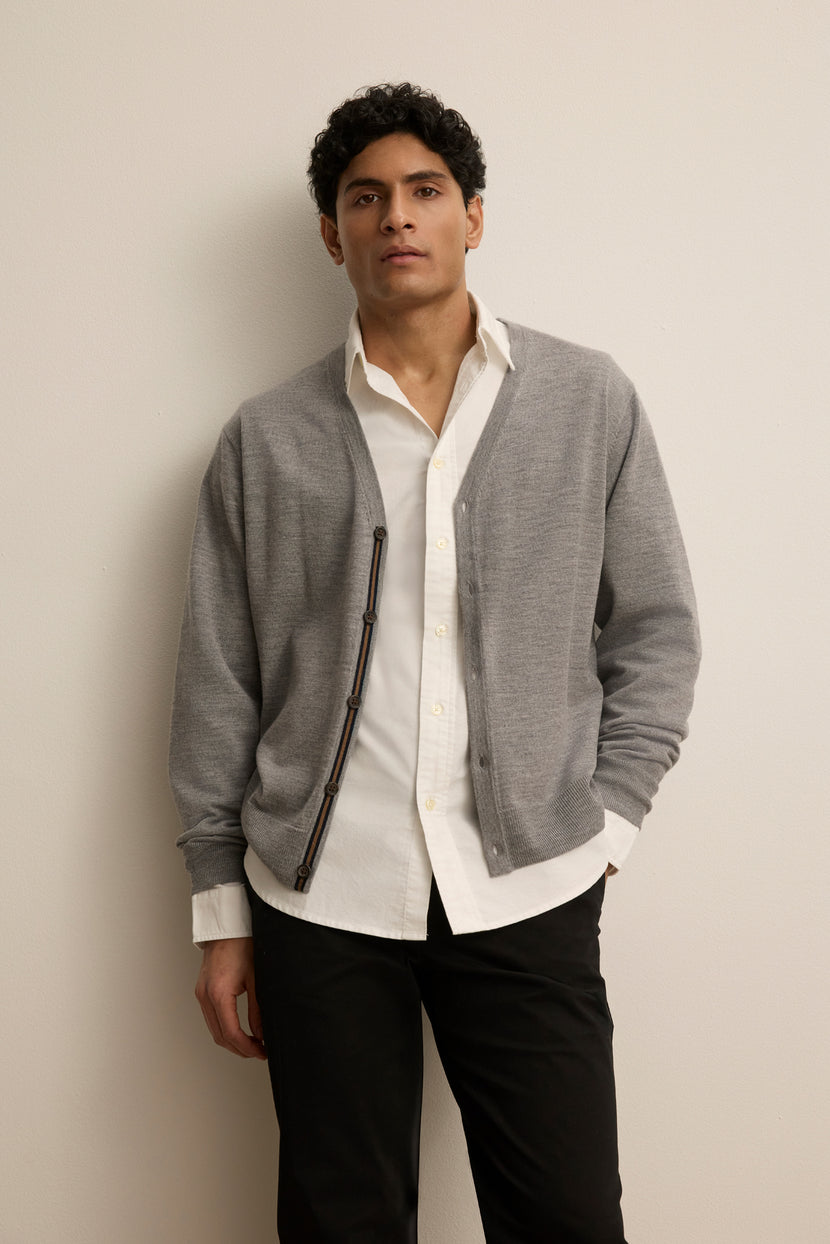 A person stands against a plain wall wearing the SILVIO CARDIGAN by Velvet by Graham & Spencer over a white button-up shirt and black pants, with one hand in their pocket.
