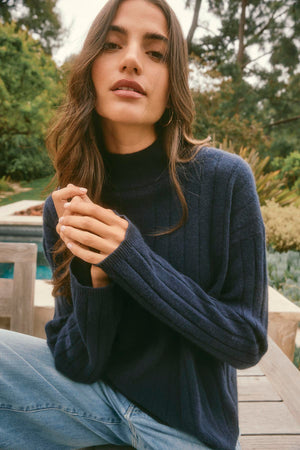 A woman in a cozy SIMONE CASHMERE MOCK NECK SWEATER from Velvet by Graham & Spencer sits outdoors, surrounded by trees, wearing jeans.