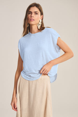 A woman stands against a neutral background wearing the Velvet by Graham & Spencer SINCLAIR TEE, styled with a beige skirt and drop earrings, effortlessly showcasing its breathable comfort.