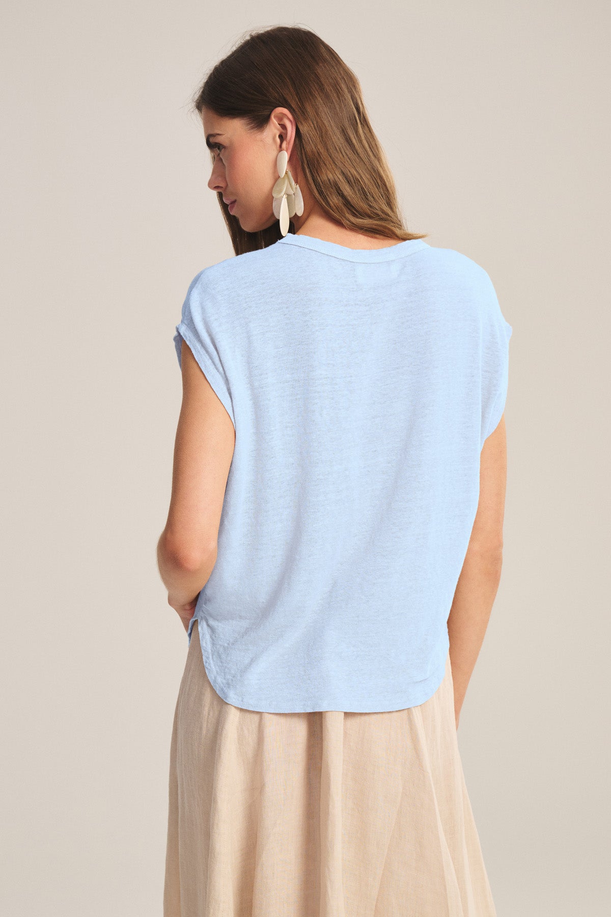 A woman with long brown hair is shown from the back in Velvet by Graham & Spencer's SINCLAIR TEE, a light blue sleeveless top, paired with a beige skirt and statement earrings for breathable comfort and effortless style.