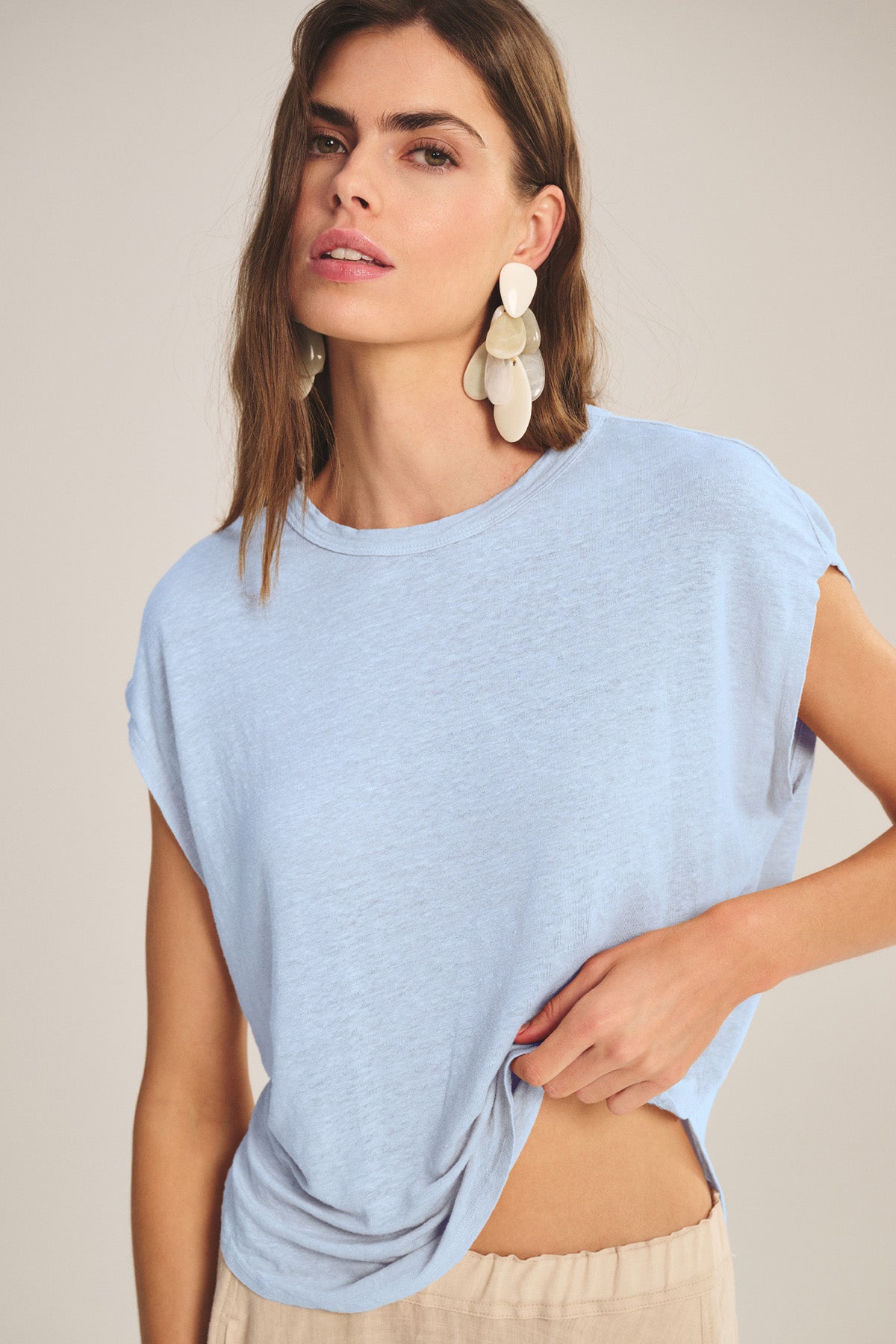 A woman wears the Velvet by Graham & Spencer SINCLAIR TEE—a light blue linen knit—paired with beige pants. With long brown hair, statement earrings, and a neutral expression, she effortlessly showcases this tee's breathable comfort and style.