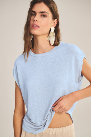 A woman wears the Velvet by Graham & Spencer SINCLAIR TEE—a light blue linen knit—paired with beige pants. With long brown hair, statement earrings, and a neutral expression, she effortlessly showcases this tee's breathable comfort and style.