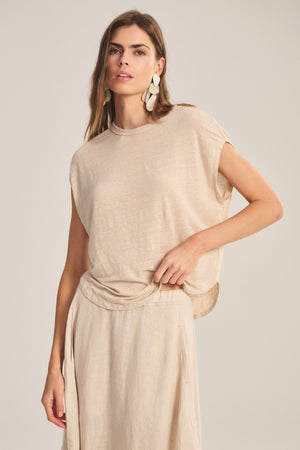 A woman wears the SINCLAIR TEE by Velvet by Graham & Spencer—a beige linen knit sleeveless top—paired with a matching skirt, showcasing breathable comfort against a plain background.