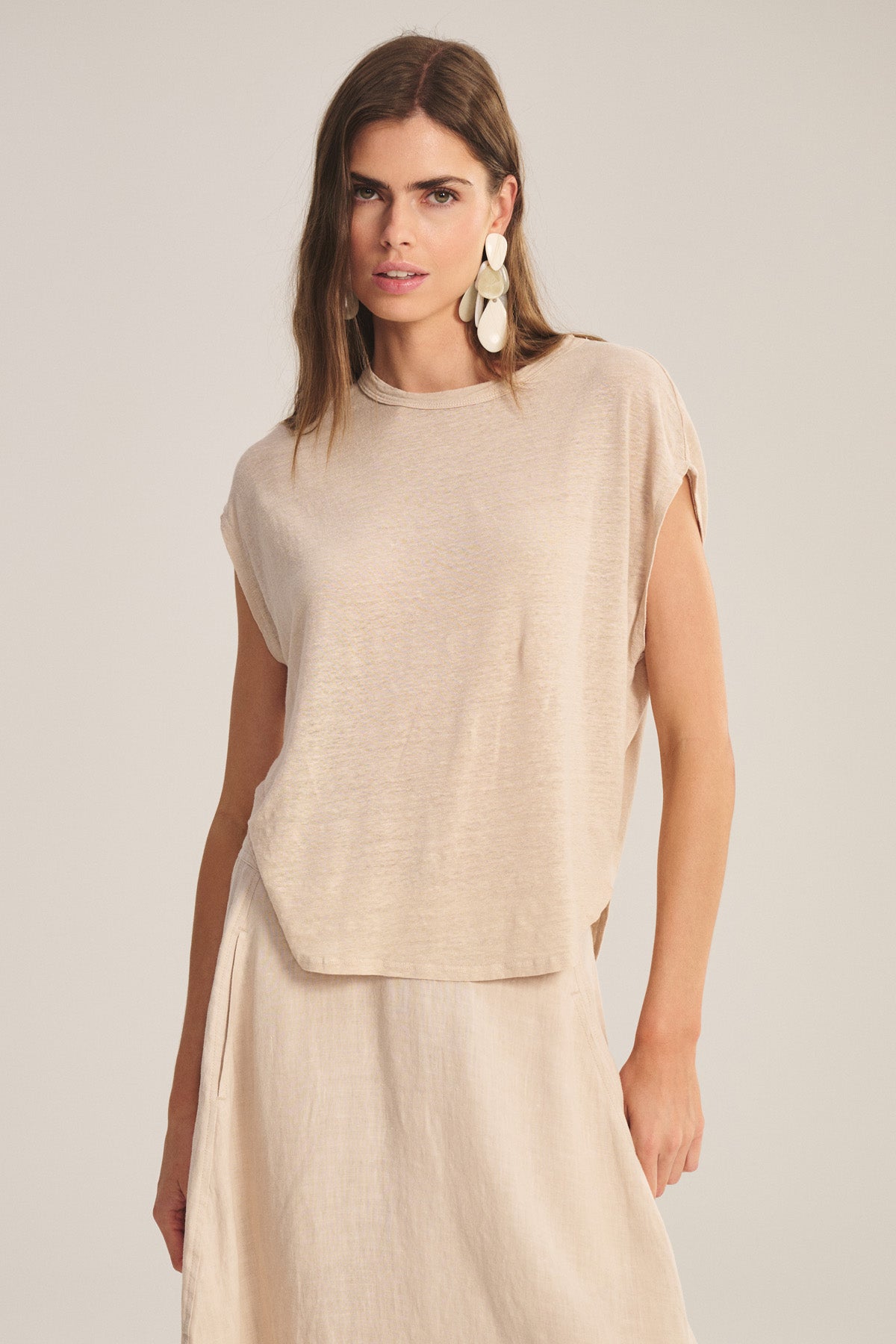 A woman models the SINCLAIR TEE by Velvet by Graham & Spencer—a loose, beige top with a cocoon silhouette—paired with a matching skirt and large white statement earrings, standing against a neutral background.
