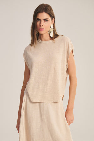 A woman models the SINCLAIR TEE by Velvet by Graham & Spencer—a loose, beige top with a cocoon silhouette—paired with a matching skirt and large white statement earrings, standing against a neutral background.