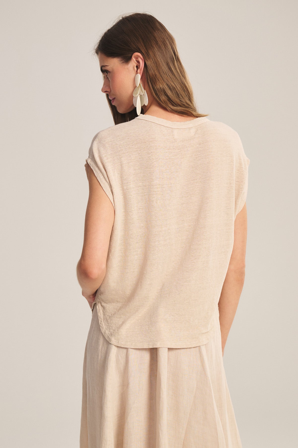 A woman with long brown hair wears the SINCLAIR TEE by Velvet by Graham & Spencer—a beige linen knit sleeveless tee—paired with a cocoon silhouette skirt and large white earrings, photographed from the back for an effortless, breathable look.