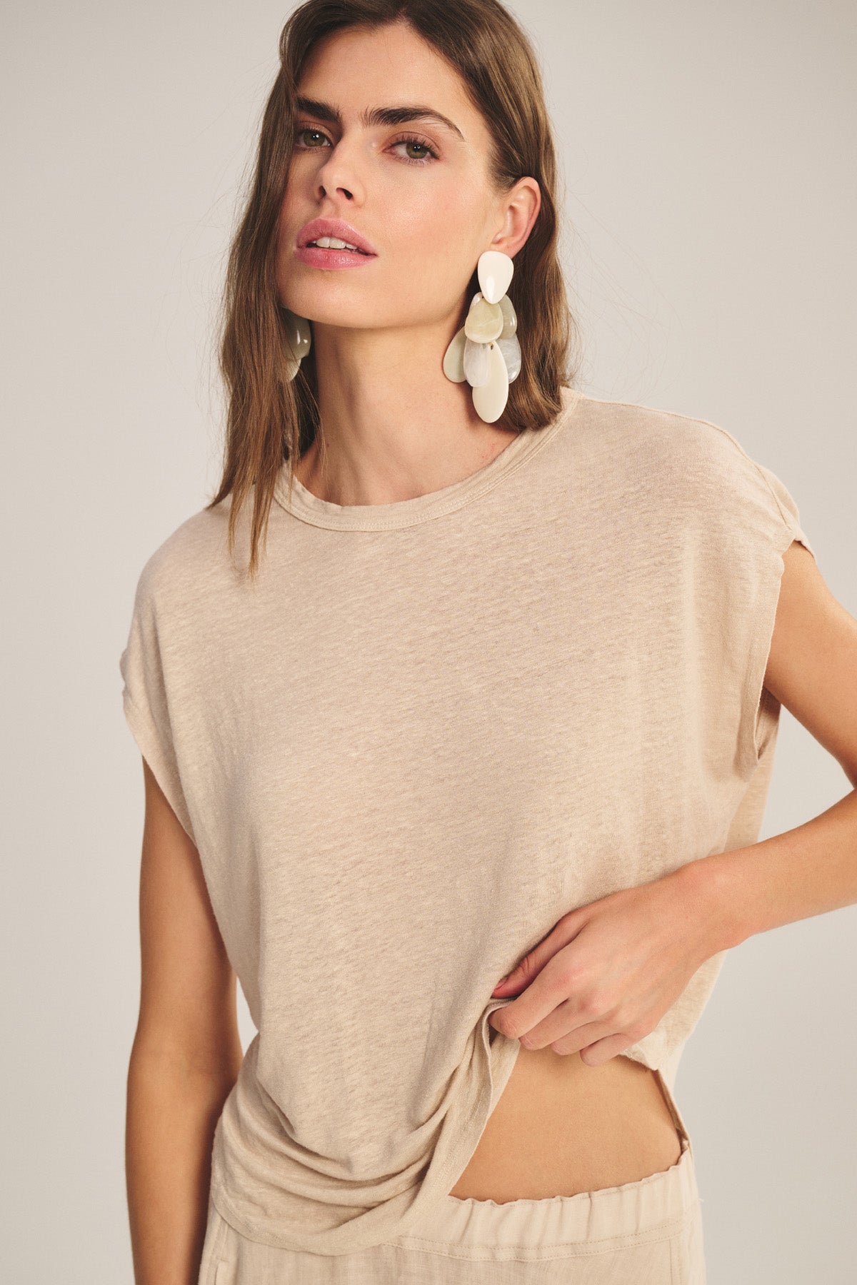A woman in the SINCLAIR TEE by Velvet by Graham & Spencer pairs its relaxed cocoon silhouette and breathable comfort with beige pants and bold white geometric earrings, gently lifting the tee to highlight its effortless style.
