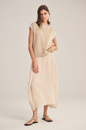 A woman wears the SINCLAIR TEE by Velvet by Graham & Spencer in beige, paired with a light beige midi skirt, black sandals, and statement earrings against a plain background.
