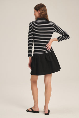 A woman faces away, wearing the SINEAD DRESS by Velvet by Graham & Spencer—a black and white striped long-sleeve dress—paired with black sandals against a plain background.