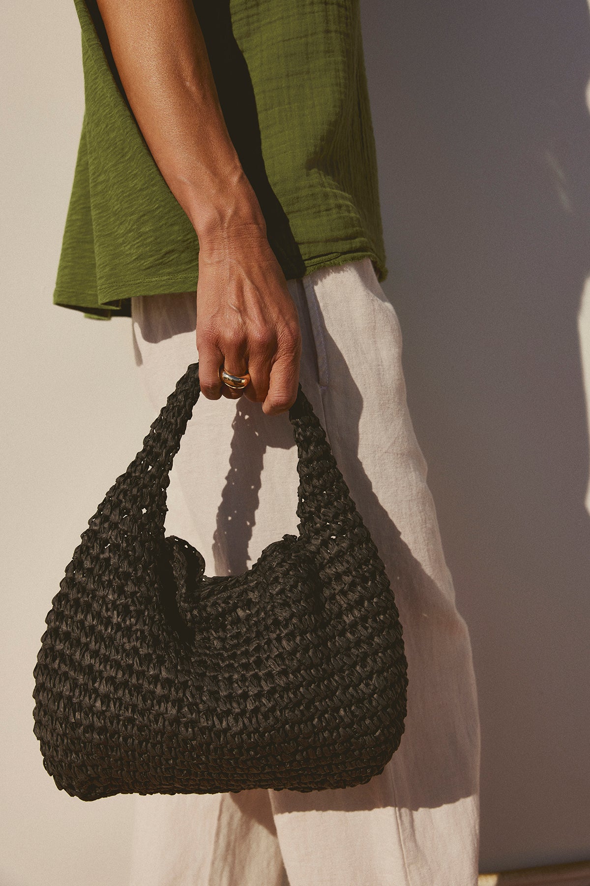   A close-up of a person holding a black Velvet by Graham & Spencer Slouch Bag, wearing a green top and white pants, with a focus on their hand and the bag. 