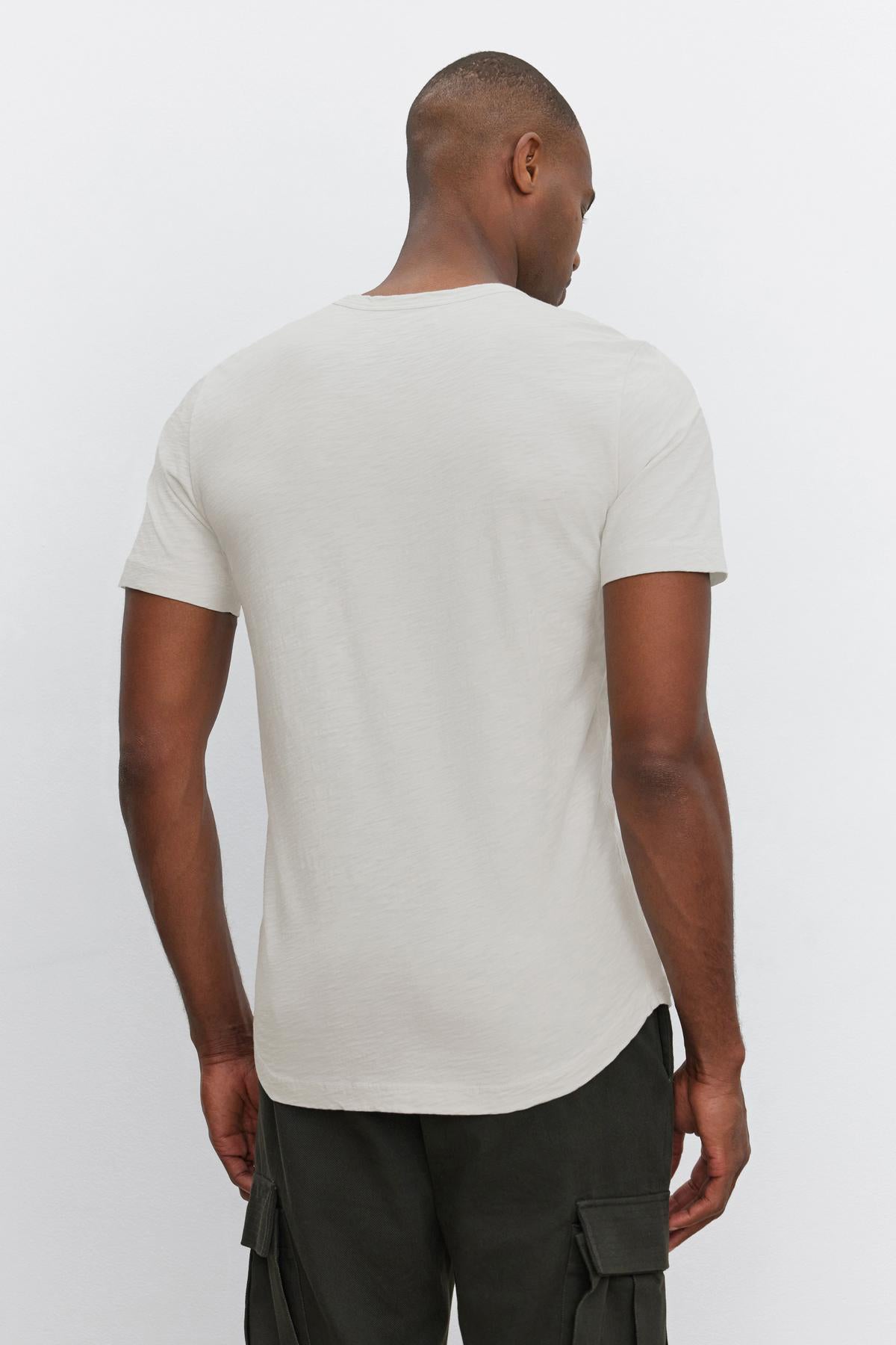   A person wearing a light gray AMARO TEE by Velvet by Graham & Spencer, featuring a classic crew neckline and short sleeves, paired with dark cargo pants, their back facing the camera. 