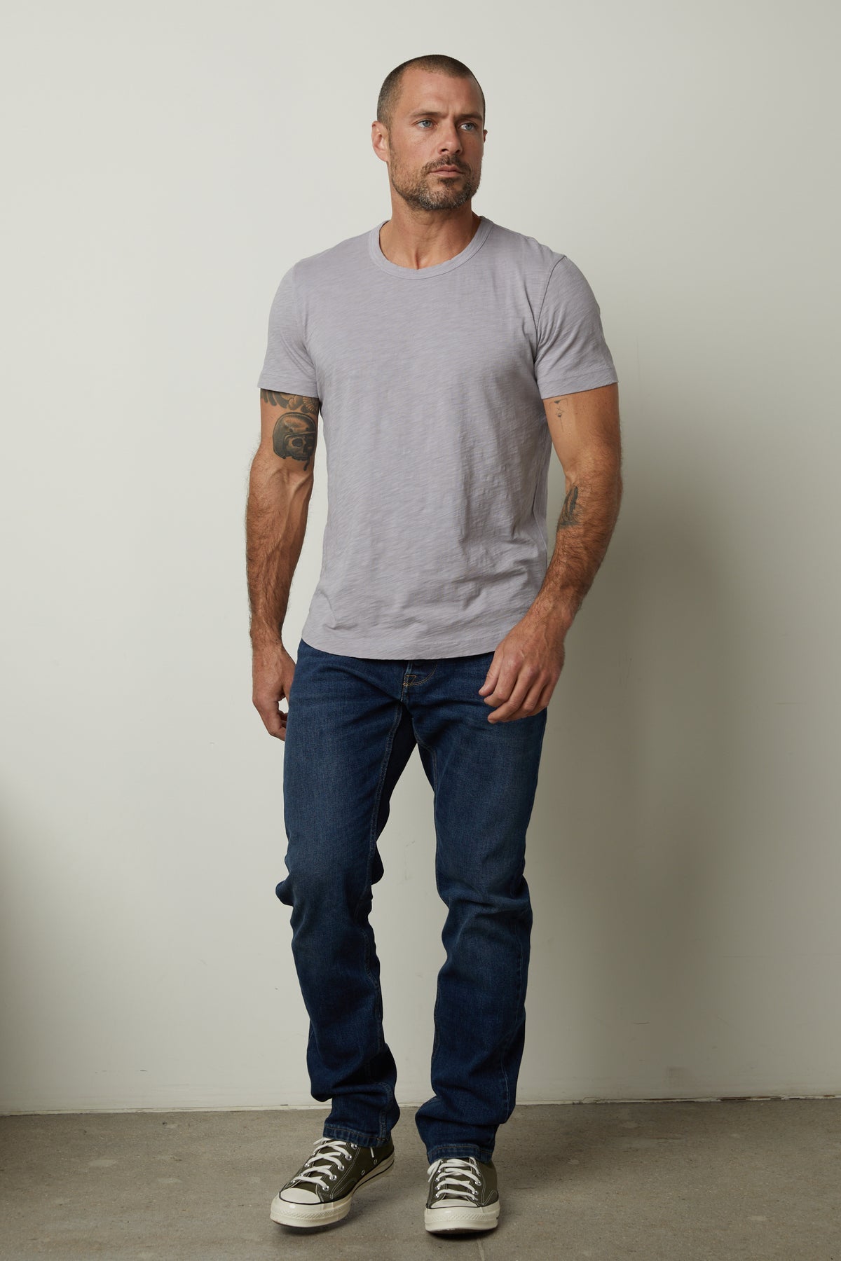   A man with short hair and tattoos on his arms stands against a plain wall, wearing a light gray, relaxed fit Velvet by Graham & Spencer AMARO TEE, blue jeans, and gray sneakers. 