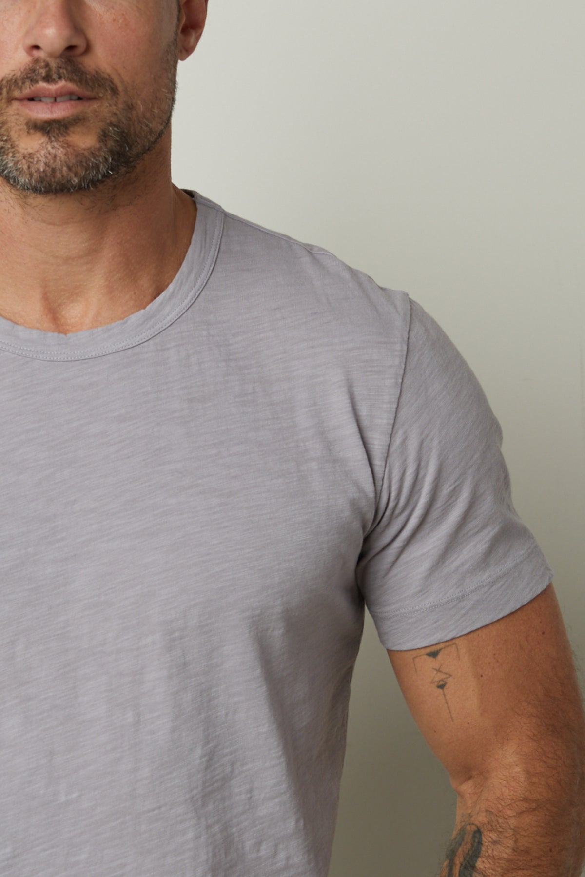   A man with a trimmed beard is wearing a light gray, cotton slub knit AMARO TEE by Velvet by Graham & Spencer, standing against a plain background. His left arm displays a tattoo. 