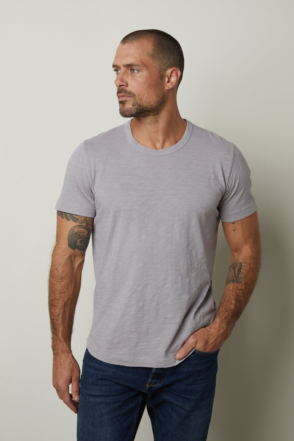 A person with short hair and a beard stands against a plain background, wearing a gray AMARO TEE by Velvet by Graham & Spencer and jeans, resting one hand in their pocket, and displaying tattoos on both arms.-37229768933569