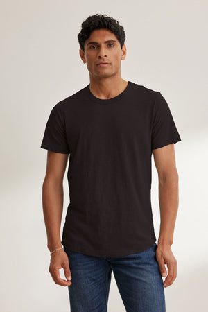 A dark-haired man stands against a light background wearing the AMARO TEE by Velvet by Graham & Spencer, made from soft Peruvian cotton. This plain black T-shirt features a classic crew neckline, perfect for layering. He's also wearing blue jeans.