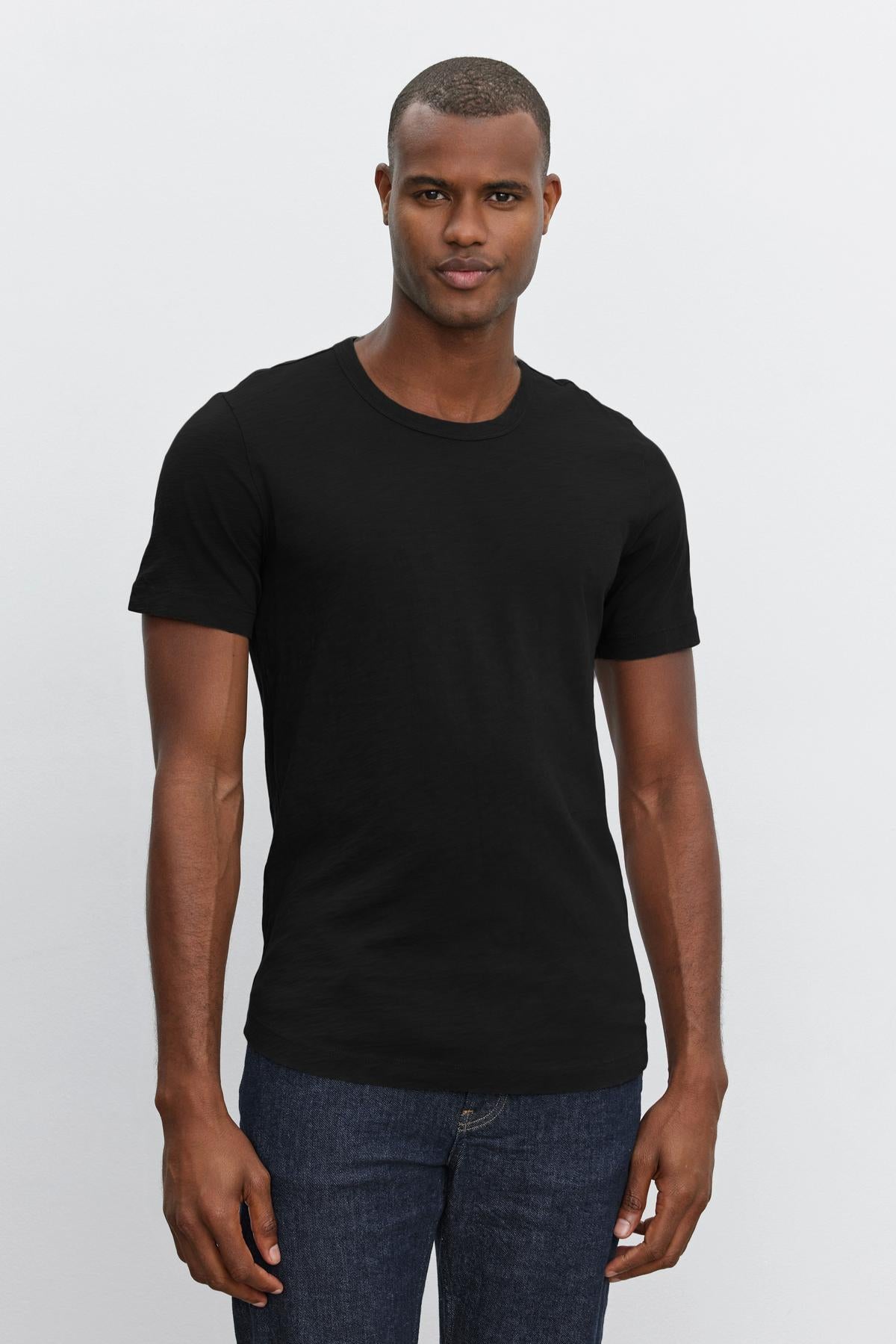 A man wearing a black Velvet by Graham & Spencer crew neck AMARO TEE and blue jeans standing against a white background.-36273921982657