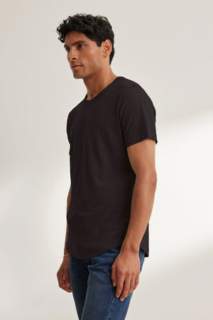The individual wearing the AMARO TEE by Velvet by Graham & Spencer, styled as a classic crew-neck black T-shirt, pairs it with jeans while standing against a plain backdrop, gazing to the side.