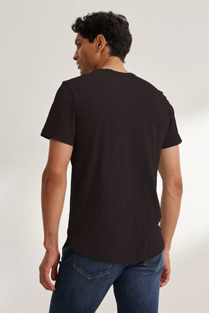 In a well-lit room, someone faces away wearing the AMARO TEE by Velvet by Graham & Spencer and jeans. The black T-shirt, crafted from soft Peruvian cotton, features a classic crew neckline, making it an excellent layering piece for any wardrobe.