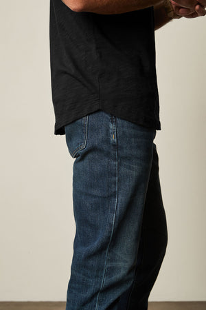 A person wearing an AMARO TEE by Velvet by Graham & Spencer in black, featuring a classic crew neckline and paired with blue jeans, stands sideways with crossed arms. The Peruvian cotton fabric makes the shirt perfect for layering in any season.