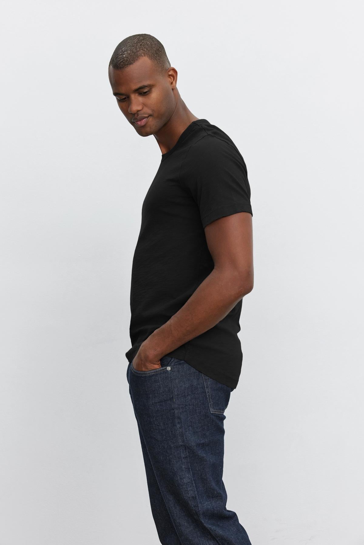 Man in a black crew neck AMARO TEE with heathered texture and jeans standing sideways against a white background by Velvet by Graham & Spencer.-36273922048193