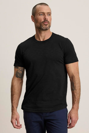 A man with short hair and a beard wears the AMARO TEE, a lightweight black Peruvian cotton T-shirt by Velvet by Graham & Spencer, paired with dark pants. Featuring a classic crew neckline, it’s perfect for layering as he faces slightly to the side against a plain background.