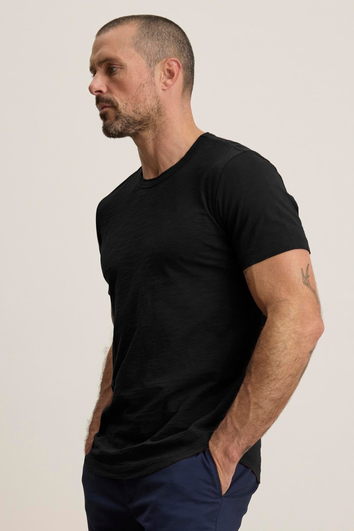   A bearded man with short hair wears the AMARO TEE by Velvet by Graham & Spencer, a black t-shirt crafted from lightweight Peruvian cotton with a classic crew neck, paired with blue pants. He's standing against a plain background with his hands in his pockets. 