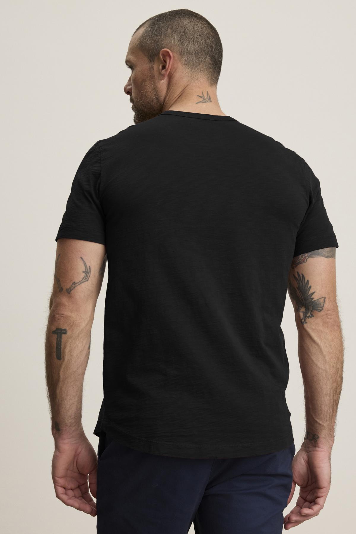 A man with tattoos, facing away, wears the AMARO TEE by Velvet by Graham & Spencer. This black t-shirt features a classic crew neckline and lightweight Peruvian cotton, pairing perfectly with dark pants against a neutral backdrop, making it an ideal layering piece.-38516739375297