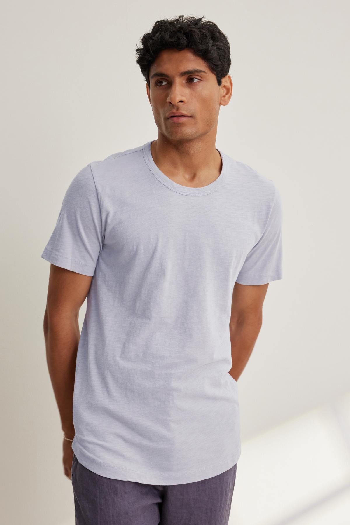   A person in the AMARO CREW NECK SLUB TEE by Velvet by Graham & Spencer, light gray and paired with dark pants, stands with hands behind their back, presenting a versatile look while gazing to the side against a plain background. 