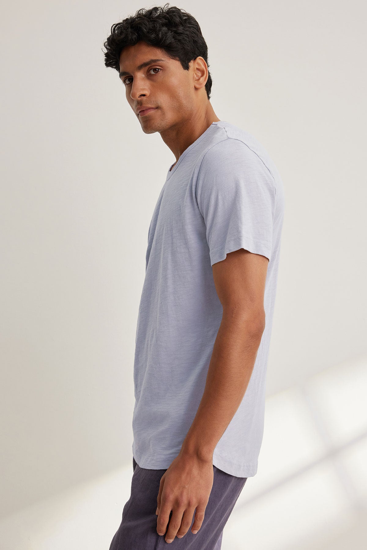 A man with dark hair, wearing an AMARO TEE from Velvet by Graham & Spencer in light gray, stands sideways in dark pants against a plain background and looks at the camera. The classic crew neck tee, crafted from soft Peruvian cotton, is perfect for layering.-38670212759745