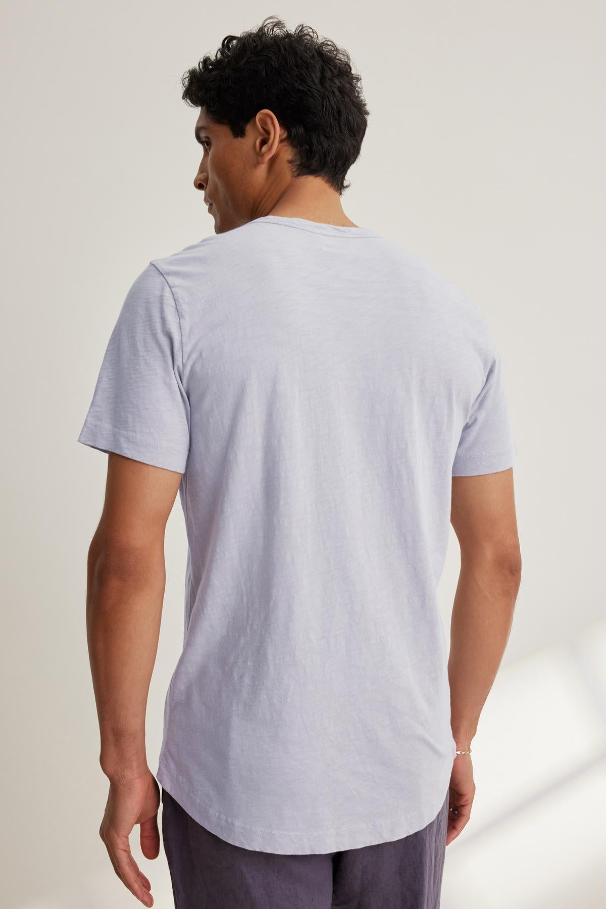   A person stands indoors in natural light, facing away to highlight a versatile look in the AMARO CREW NECK SLUB TEE by Velvet by Graham & Spencer—a light gray cotton slub T-shirt with a classic crew neckline—paired with dark pants. 