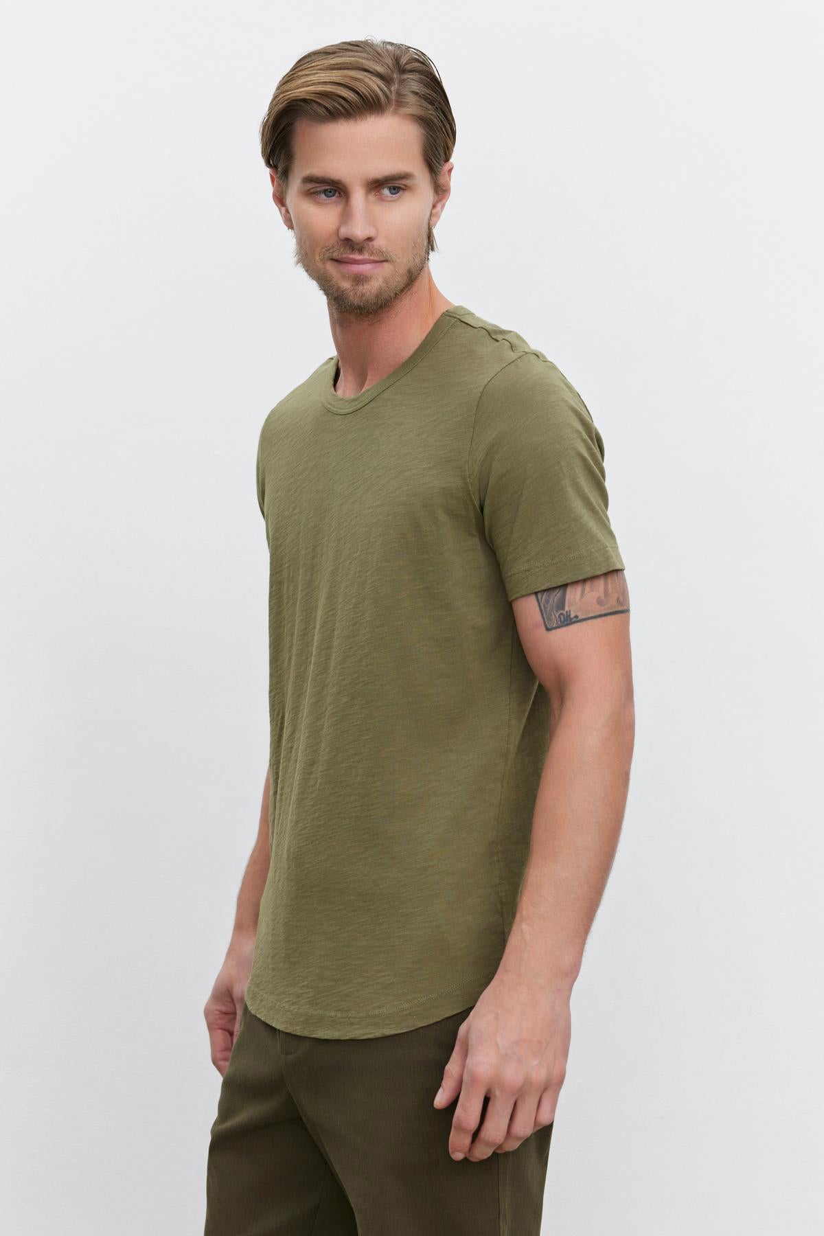 A man with light hair wearing an olive green AMARO TEE by Velvet by Graham & Spencer, made from lightweight Peruvian cotton, along with matching pants stands against a plain white background.-37682021793985