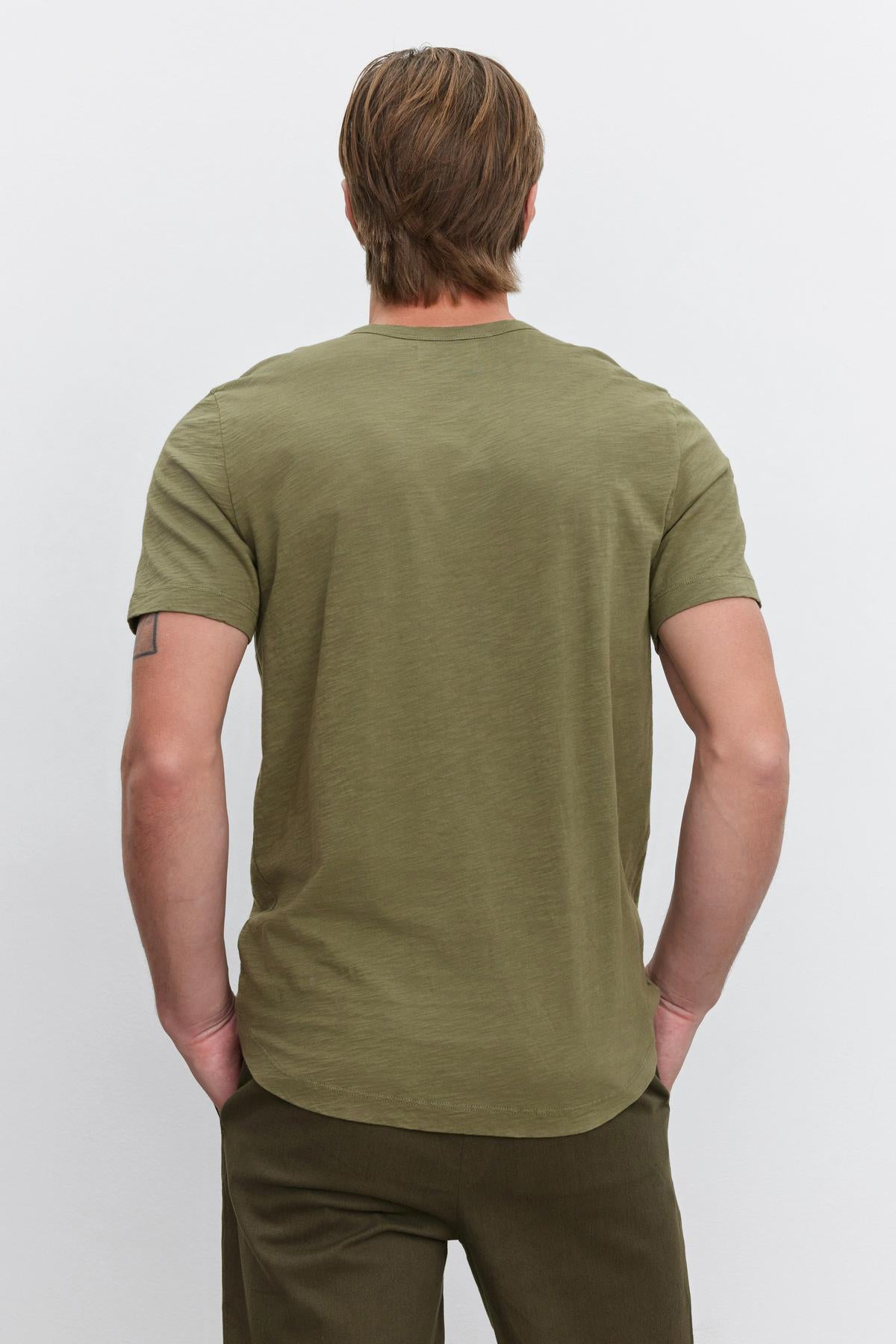   A person with light brown hair wearing an olive green AMARO TEE by Velvet by Graham & Spencer and dark green pants is standing with their back to the camera. 