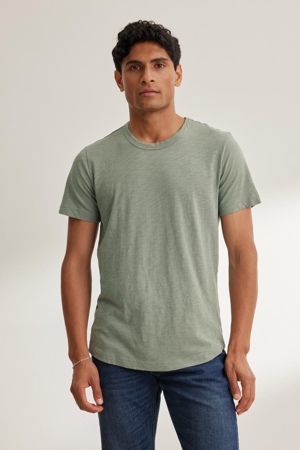   A man stands against a plain backdrop wearing the AMARO TEE by Velvet by Graham & Spencer, a light green crew neck t-shirt paired with blue jeans. Made of lightweight Peruvian cotton, it's perfect for layering in any season. 