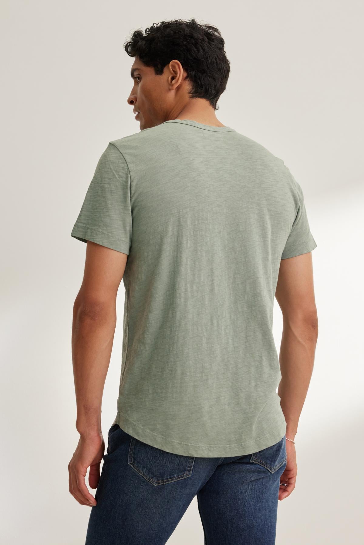   A man with short dark hair, seen from the back, wears the AMARO TEE by Velvet by Graham & Spencer. The light green T-shirt has a classic crew neckline and is made of lightweight Peruvian cotton. He pairs it with blue jeans. 