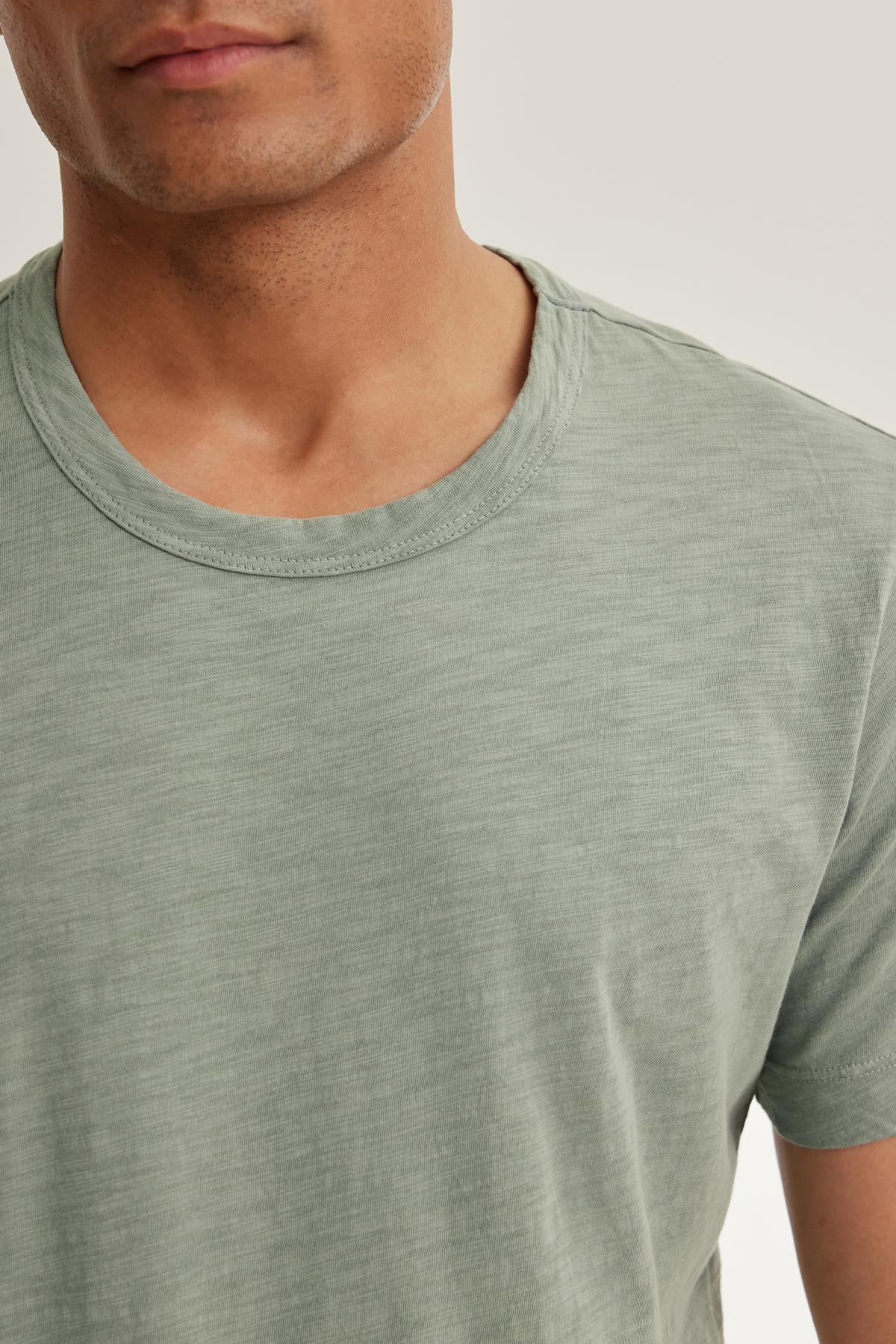   A person wears the AMARO TEE from Velvet by Graham & Spencer, a light green, classic crewneck shirt crafted from lightweight Peruvian cotton, shown from shoulders up against a neutral backdrop. The polished finish adds elegance to the simple and stylish look. 