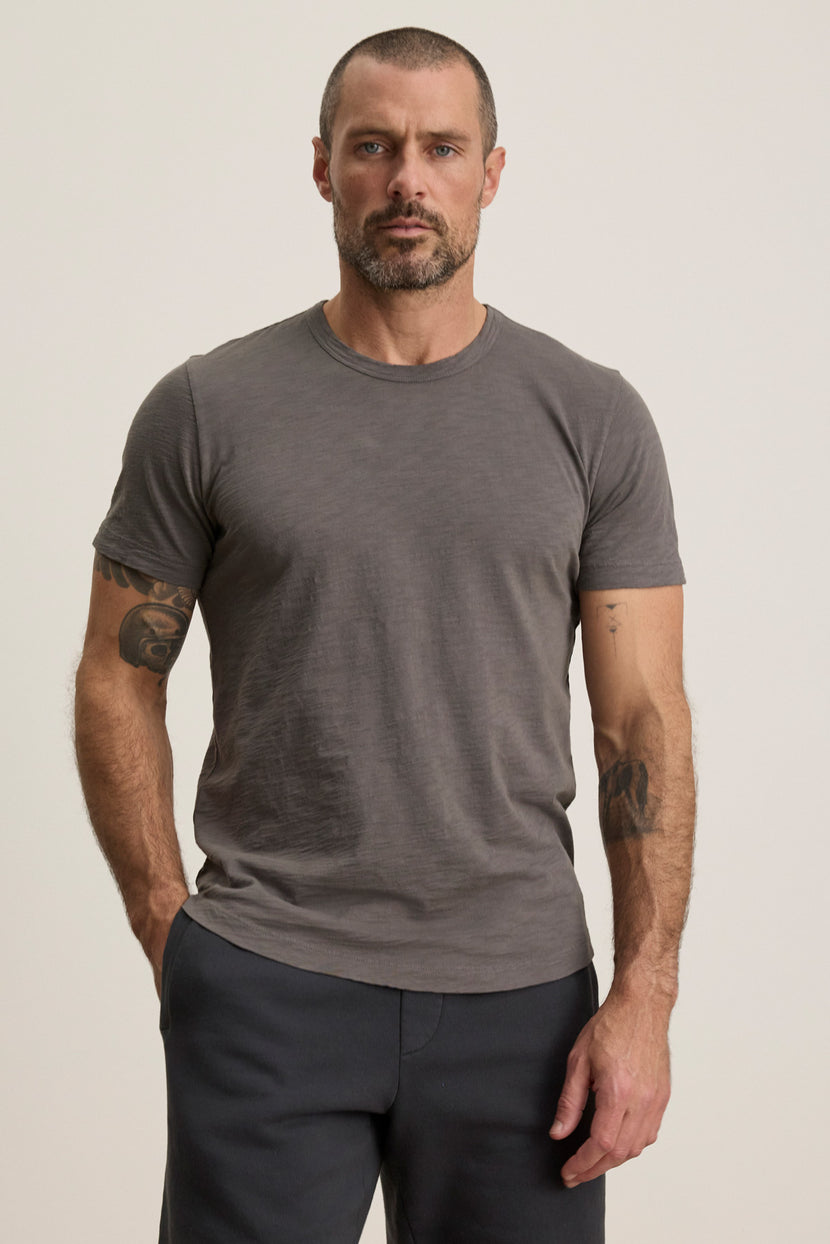 A man with short hair and a beard wears the AMARO TEE by Velvet by Graham & Spencer—a grey Peruvian cotton crewneck—paired with dark pants, standing against a light background with one hand in his pocket. A perfect layering piece.