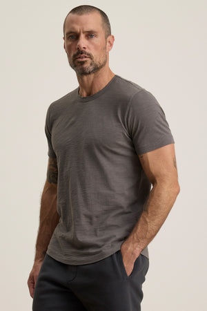 A man with short hair and a beard wears the gray AMARO TEE by Velvet by Graham & Spencer, featuring Peruvian cotton and a classic crew neckline, paired with dark pants—an ideal layering piece against a light background.