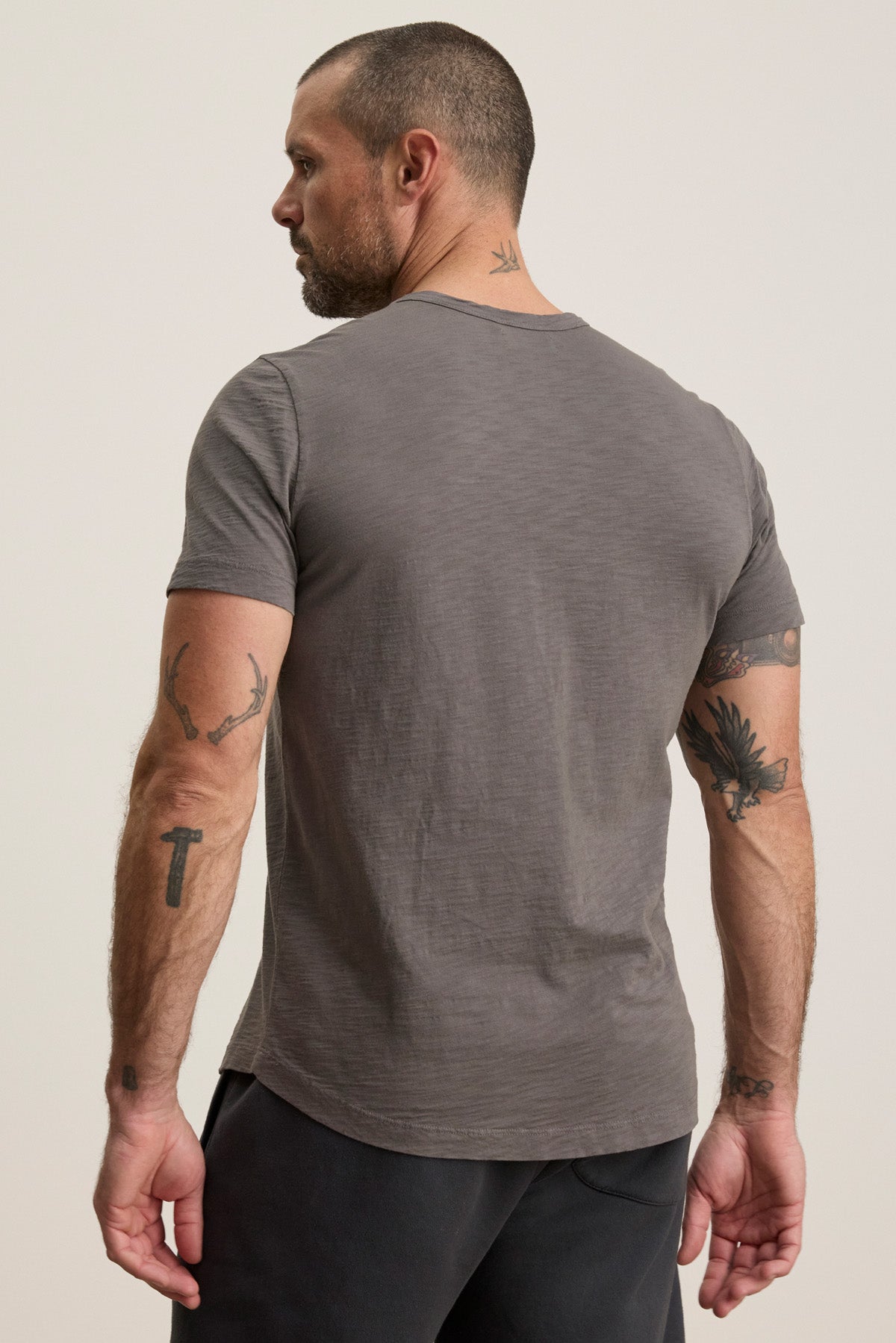 A man with short hair and tattoos stands with his back to the camera, wearing the AMARO TEE by Velvet by Graham & Spencer—a grey Peruvian cotton crewneck t-shirt, perfect as an everyday layering piece.-40452464836801