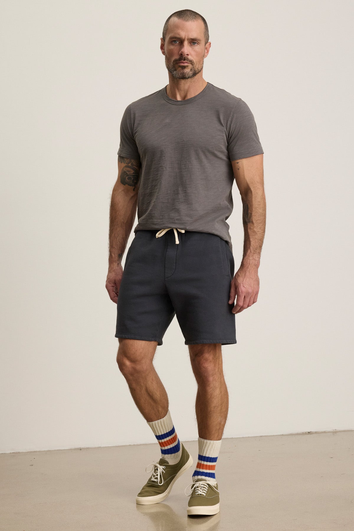 A man stands indoors wearing the AMARO TEE by Velvet by Graham & Spencer in grey Peruvian cotton with a crew neckline, paired with dark drawstring shorts, striped socks, and olive green sneakers.-40452464738497