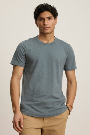A person with short curly hair wears the AMARO TEE by Velvet by Graham & Spencer, a blue crew neck t-shirt, and tan pants, standing against a plain light background with one hand in their pocket.