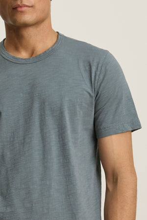 A person is shown from shoulders to waist, facing forward, wearing the Velvet by Graham & Spencer AMARO TEE in light blue with a short sleeve and crew neckline; their face is partially out of frame.