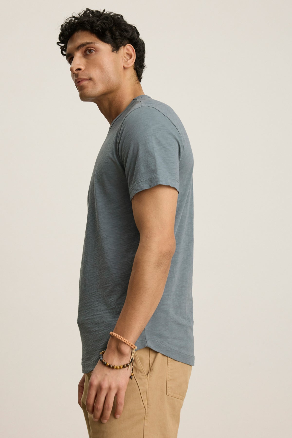 A man stands sideways against a plain background wearing the AMARO TEE by Velvet by Graham & Spencer, a blue lightweight cotton crewneck, paired with tan pants and a beaded bracelet.-39446016098497