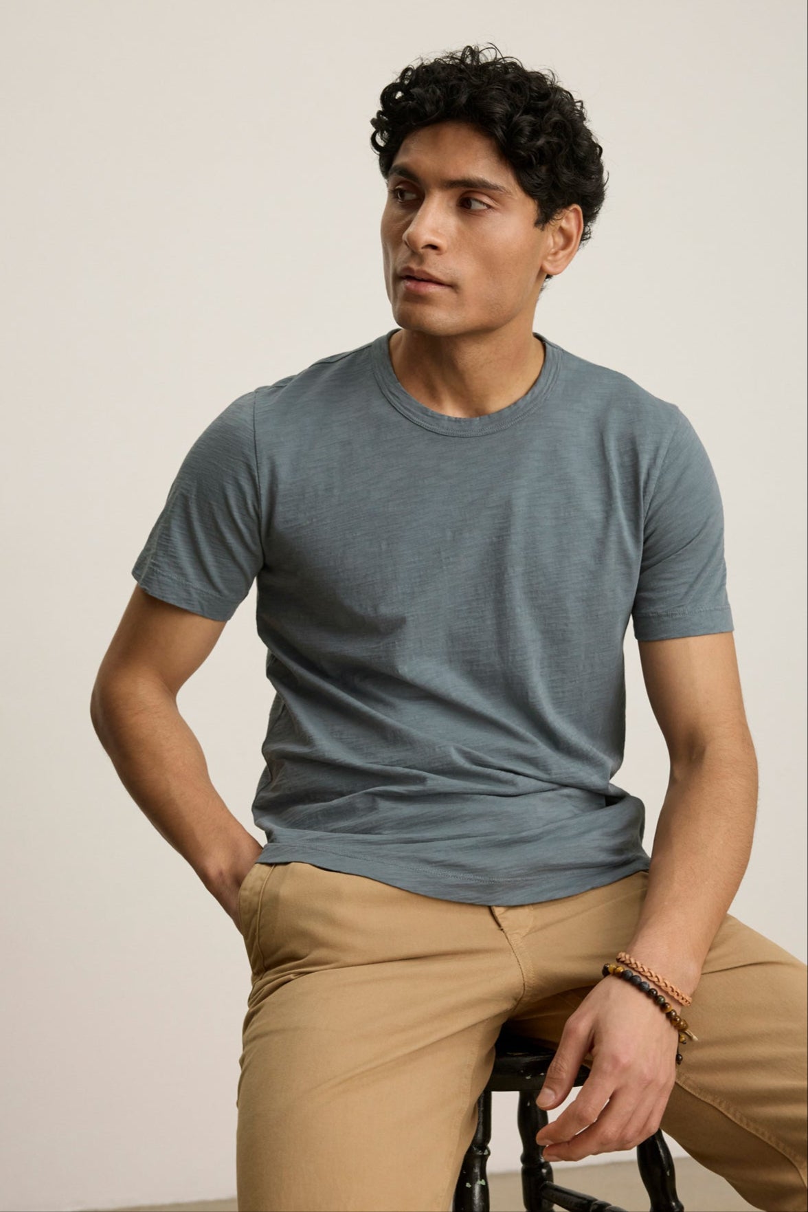 A man with short curly hair wears the AMARO TEE by Velvet by Graham & Spencer and beige pants, sitting on a stool against a plain light background.-39446016164033