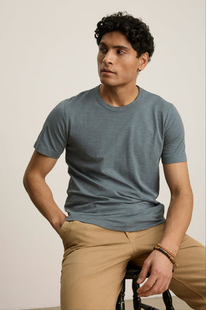 A man with short curly hair wears the AMARO TEE by Velvet by Graham & Spencer and beige pants, sitting on a stool against a plain light background.